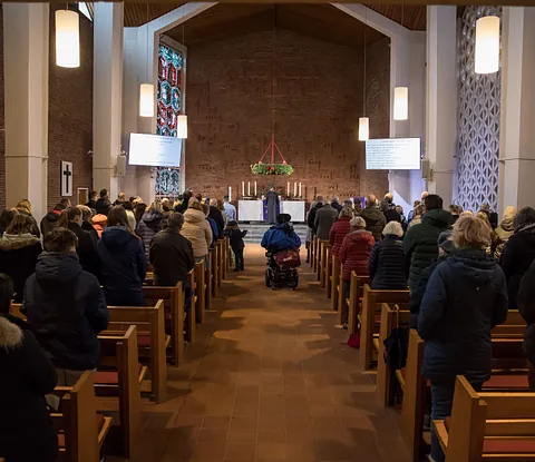 Innenraum der Paulskirche in der totale. Im Zentrum befindet sich der Altar mit sech Kerzen. Darüber ein Adventskranz mit brennenden Kerzen. Links von Altar die Taufkerze und ein Buntglasfenster. Ein Gottesdienst findet statt, die Feiernden stehen.